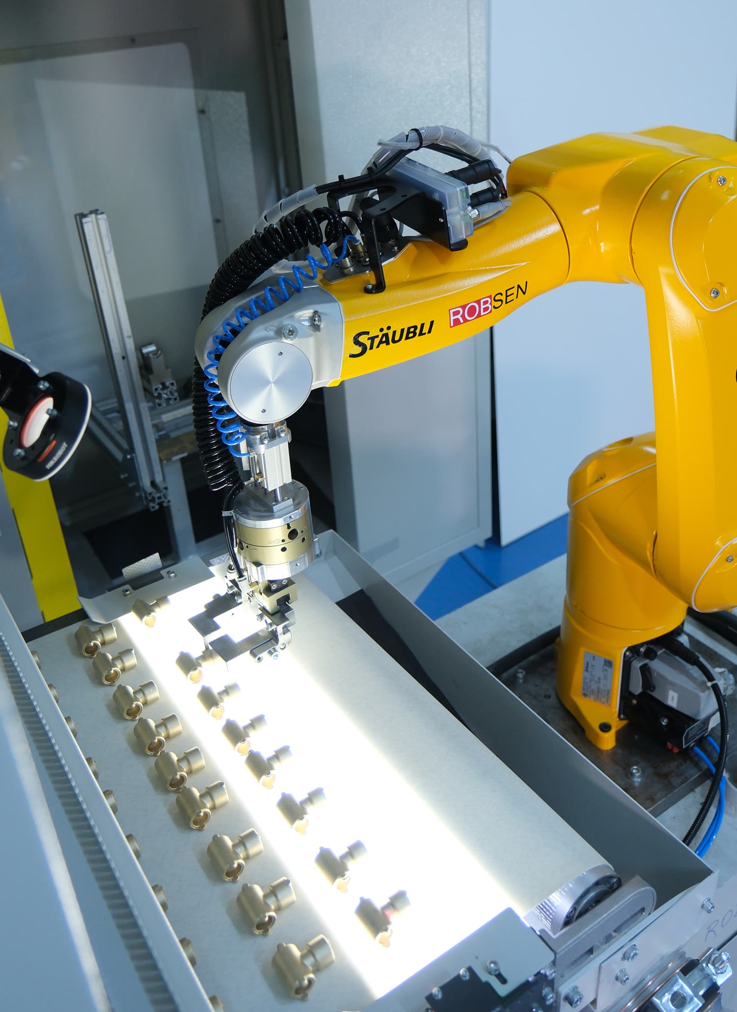 Yellow Stäubli robot using a compensating gripper to pick forged metal parts from the illuminated conveyor of an IROCUBE MS60L cell.